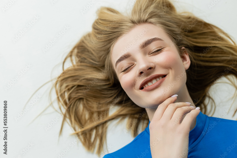 Fototapeta premium top view of happy blonde woman lying and touching chin on grey background.