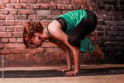 A young slender girl in black leggings and a birch T-shirt sits in an asana against the background of a brick wall.