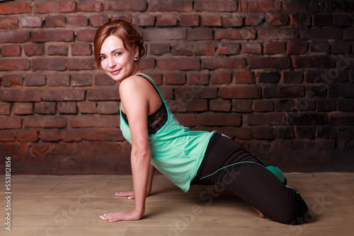 A young slender girl in black leggings and a birch T-shirt sits in an asana against the background of a brick wall.