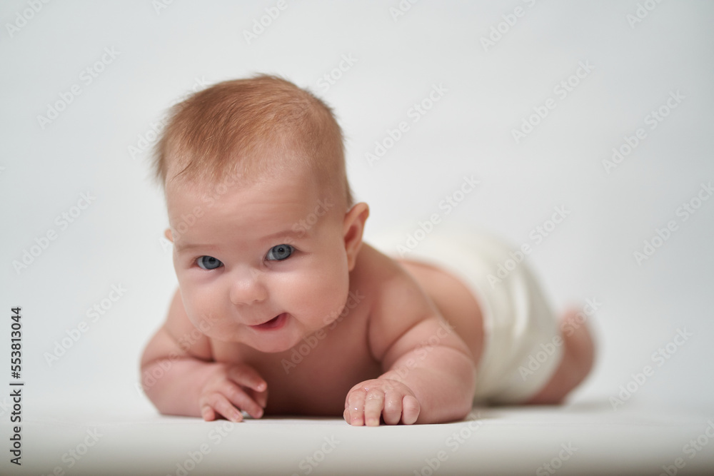 The baby is lying on his belly, slyly looking at the camera against a white background. The baby is lying on his belly, slyly looking at the camera against a white background.