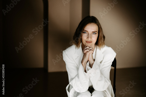 Portrait of successful woman, with natural makeup with face, wearing in white jacket and holding hands near chin, posing in dark studio with beige walls