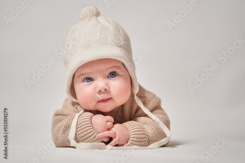 A portrait of an infant in knitted clothes and a white cap looks at the camera