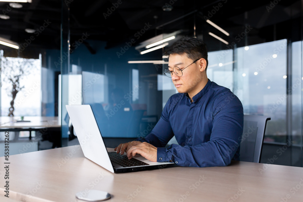 Serious and thoughtful businessman working inside office sitting at table using laptop at work, mature asian boss in shirt thinking and typing on keyboard. Serious and thoughtful businessman working inside office sitting at table using laptop at work, mature asian boss in shirt thinking and typing on keyboard.