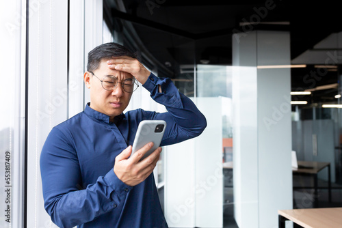Upset asian man in office at work reading bad news from phone online, businessman in shirt near window working inside office, using smartphone.