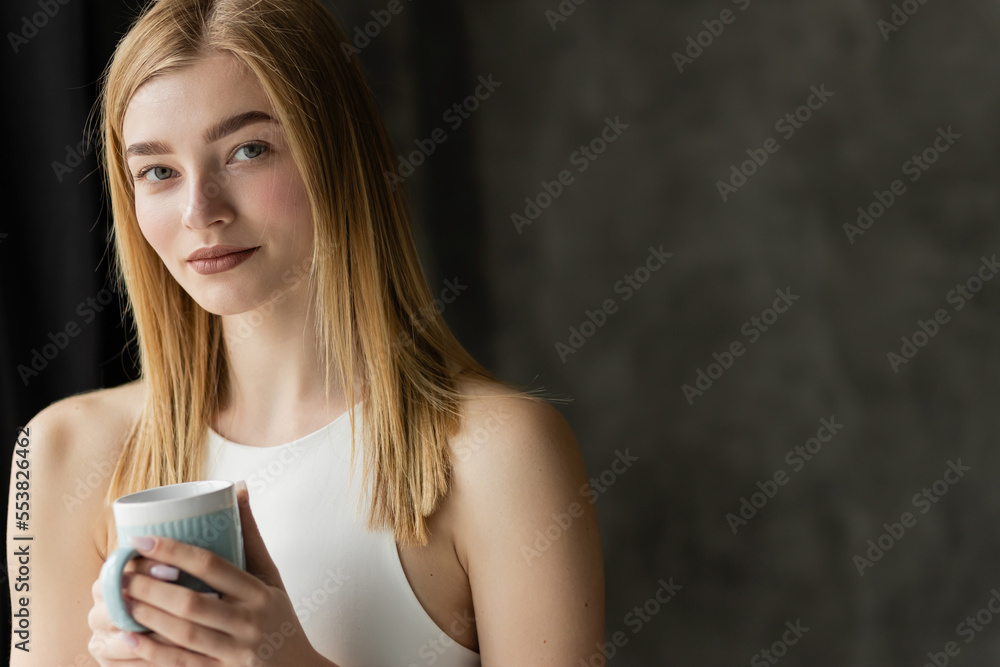 Fototapeta premium Blonde woman in top holding cup and looking at camera at home.