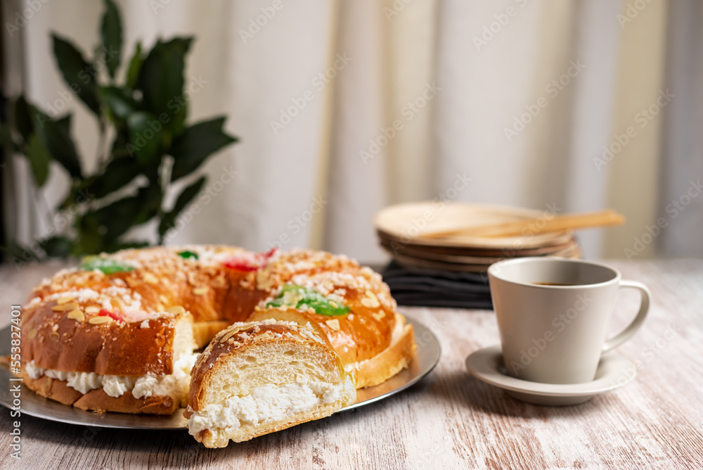 Roscon de reyes with a cut piece revealing the cream of its filling, along with a cup of coffee, in the background several wooden serving dishes. Roscon de reyes with a cut piece revealing the cream of its filling, along with a cup of coffee, in the background several wooden serving dishes.
