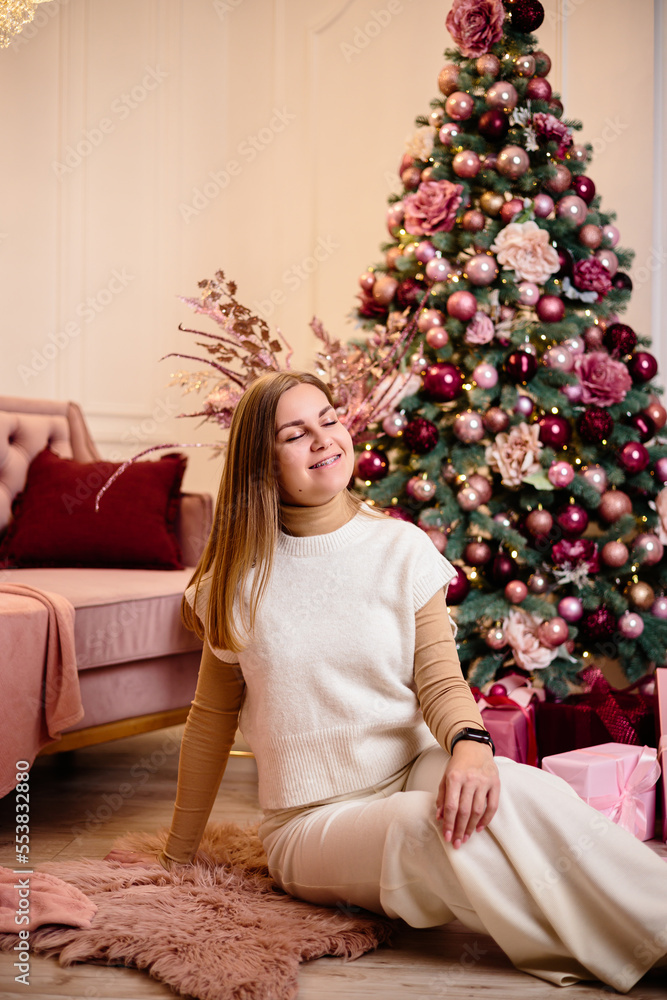A stylish happy young woman in a fashionable white knitted suit is sitting on a soft rug in a cozy room near a Christmas tree. Fashionable girl joyful model rests in the Christmas studio