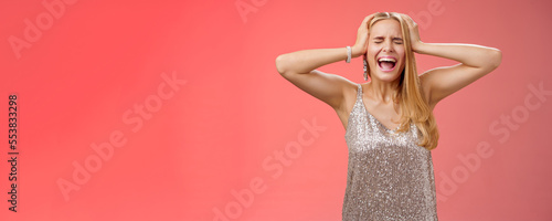 Upset distressed pressured fed up panicking young blond woman in dress scream cry heart out being heartbroken close eyes hold hands head mentaly unstable standing sad depressed red background photo