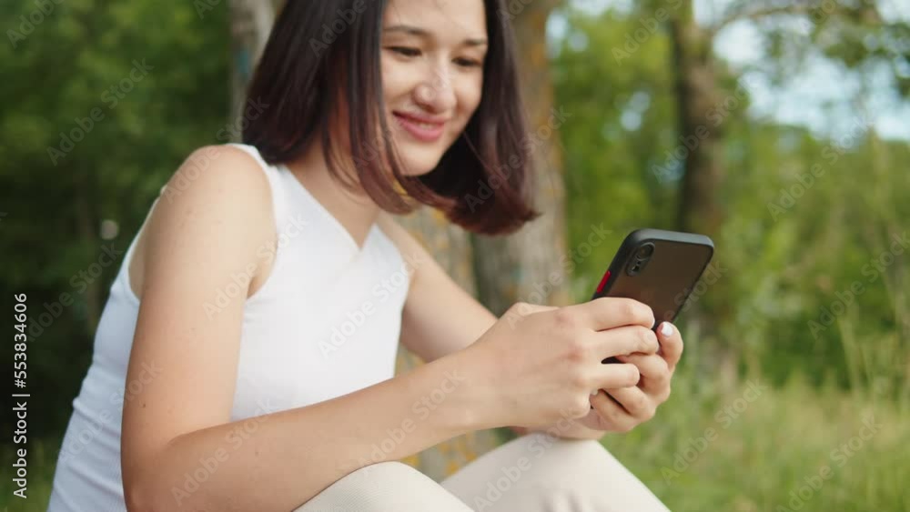 Woman using phone outdoor. Young student holding smartphone, cheerful female person sitting in park and smiling. Remote work, freelance. Sunny summer day. Woman using phone outdoor. Young student holding smartphone, cheerful female person sitting in park and smiling. Remote work, freelance. Sunny summer day.