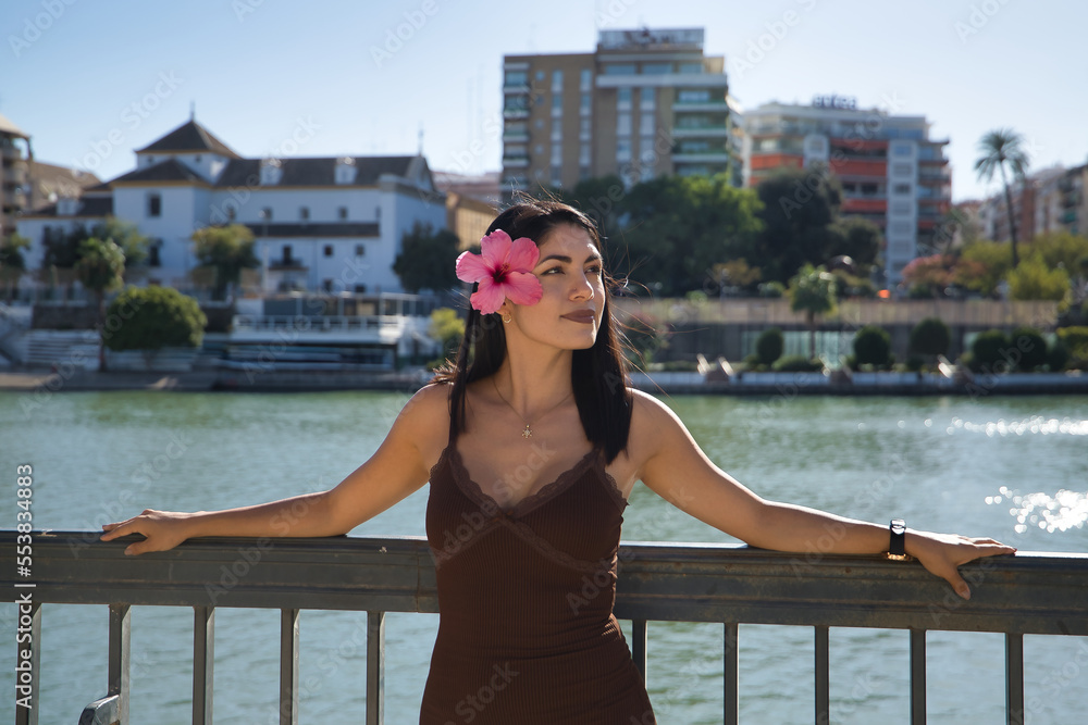 South American woman, young and beautiful, brunette, in brown dress, leaning on a railing with a beautiful flower in her ear. Concept fashion, beauty, flowers, travel, holidays, relaxation.