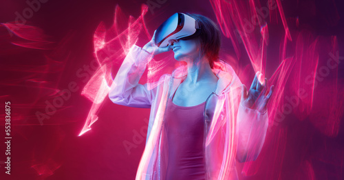 Woman in futuristic costume. Girl in glasses of virtual reality while touching air. Augmented reality game  future technology  AI concept. VR. Neon red light. Viva magenta color of the year 2023. 