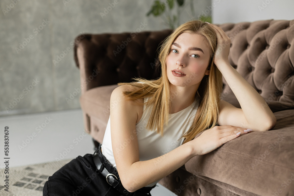 Fototapeta premium Portrait of young blonde woman looking at camera near couch at home.