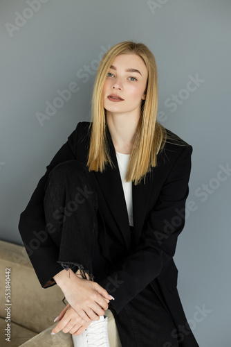 Portrait of blonde woman in black jacket looking at camera near couch at home.