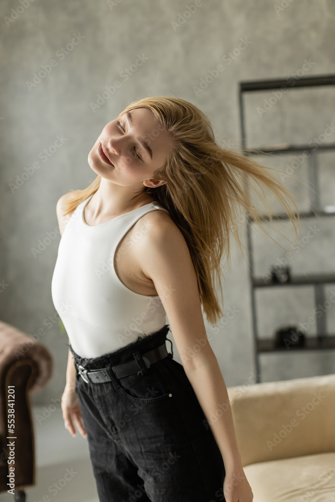pretty blonde woman in white tank top dancing with closed eyes at home.
