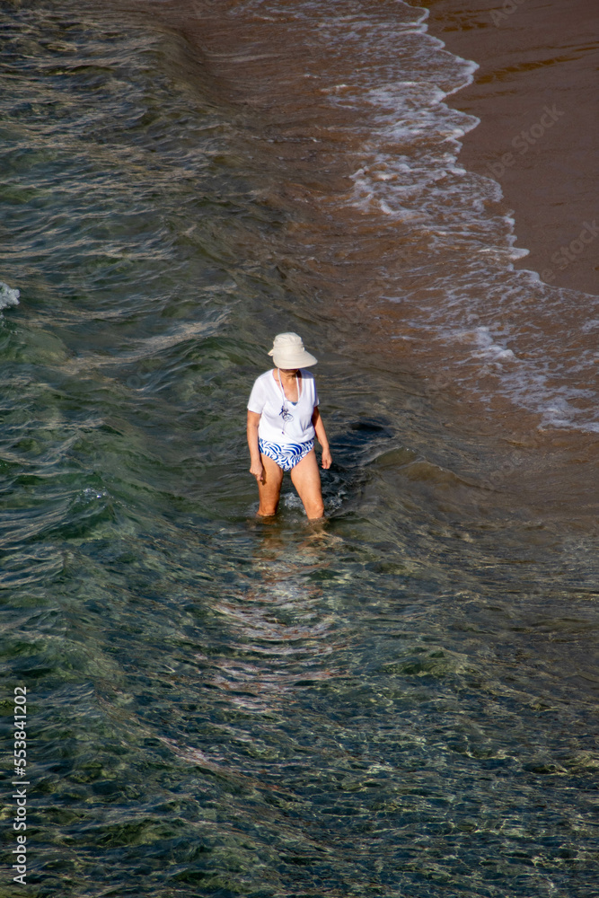 Mujer de raza caucásica turista caminando por la orilla de la playa de Benidorm con una gorra para protegerse del sol.