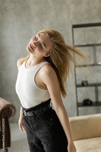 pretty blonde woman in white tank top dancing with closed eyes at home.