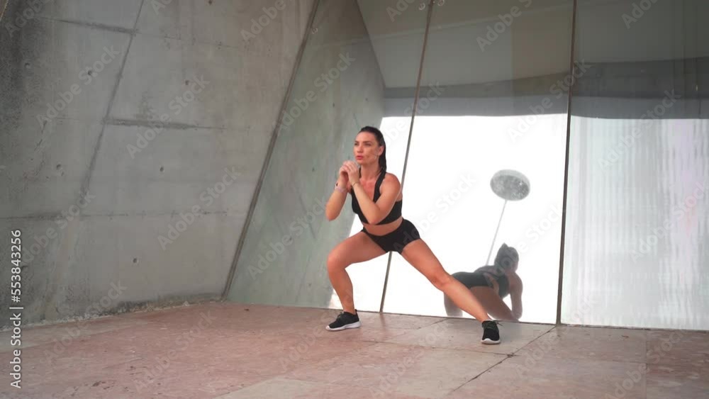 Fit middle age woman doing side lunges exercising outdoors. Female athlete warming up muscles and stretching while standing near mirror wall. Confidence sporty female training alone Fit middle age woman doing side lunges exercising outdoors. Female athlete warming up muscles and stretching while standing near mirror wall. Confidence sporty female training alone