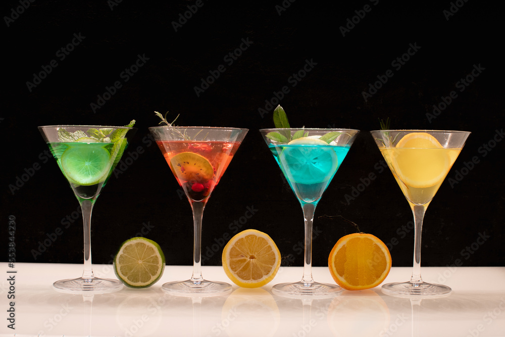 Quatre verres de différents cocktail avec des cocktails colorés sur un fond noir avec des reflets de fruits sur le comptoir.	