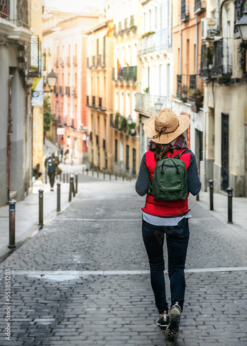 A female tourist with her back turned, wearing a hat and backpack, walks down a street in the historic center of Madrid. Hiking tourism in Spain. Concept of Spanish tourism and culture.