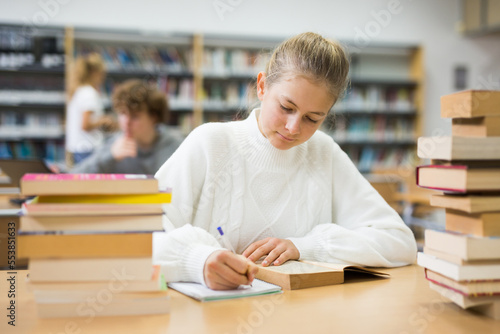 Teenage girl reading books and doing homework in library.