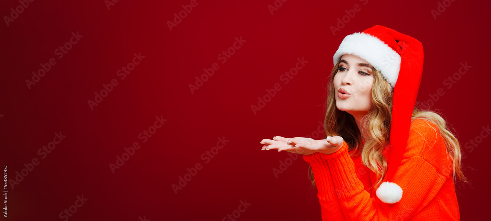 banner. a woman in a Santa Claus hat sends an air kiss on a red background. banner. a woman in a Santa Claus hat sends an air kiss on a red background.