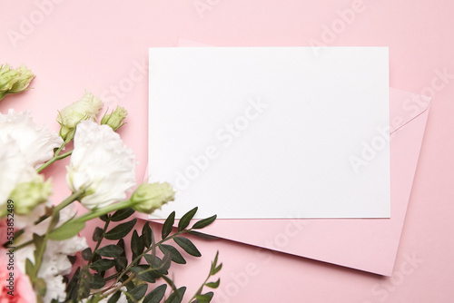 Holiday greeting card mockup with envelope and white flowers on pink background, top view, flat lay. Blank wedding invitation card mockup and floral decor