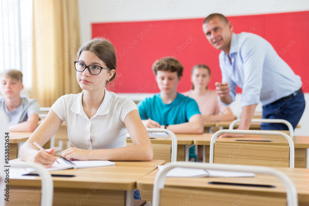 Teenager sitting in class room. Male teacher explaining something to them.