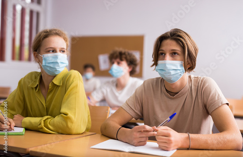 Smart teenager in protective mask studying in classroom, listening to lecturer and writing in notebook