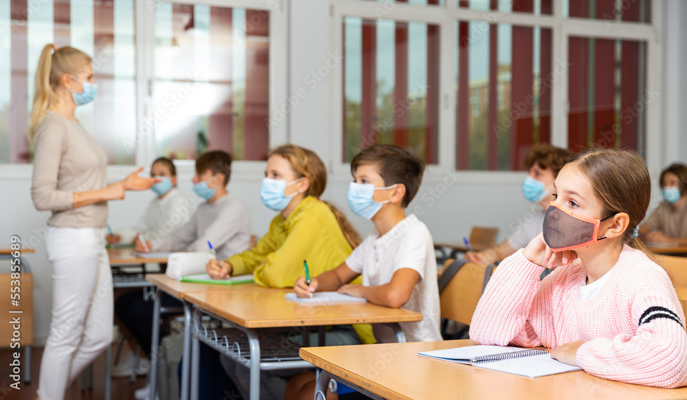 Smart teenager in protective mask studying in classroom, listening to lecturer and writing in notebook