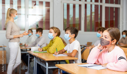 Smart teenager in protective mask studying in classroom, listening to lecturer and writing in notebook