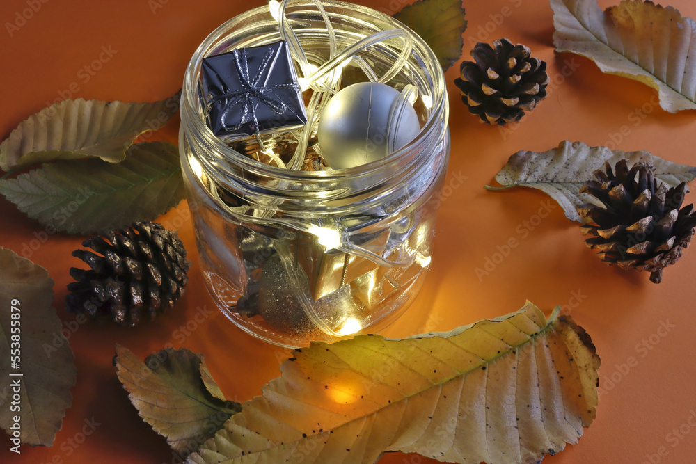 Christmas decorations and pine cones in a jar with a lighted chain on an orange background with leaves and pine cones from a tree