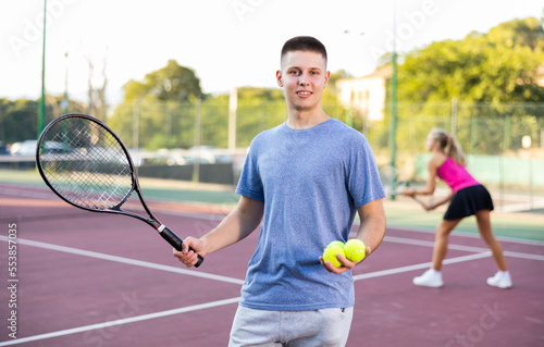 Portrait of positive young man with racket and ball in his hands on the tennis court outdoor © JackF