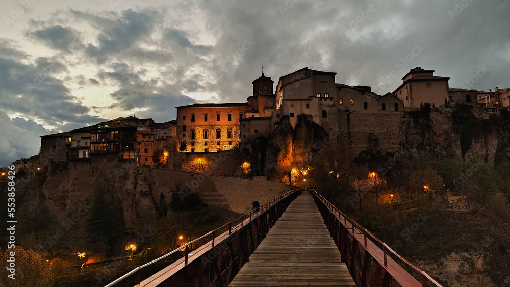 old town of Cuenca in Spain, from the bridge overlooking the medieval buildings of the city at evening with a cloudy sky in the background and orange lights - touristic postcard for a wallpaper