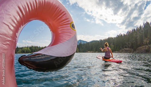 Slika na platnu Adventurous athletic woman sitting on a paddle board on a large alpine lake in the Pacific Northwest on a beautiful summer day with a blowup flamingo in the foreground
