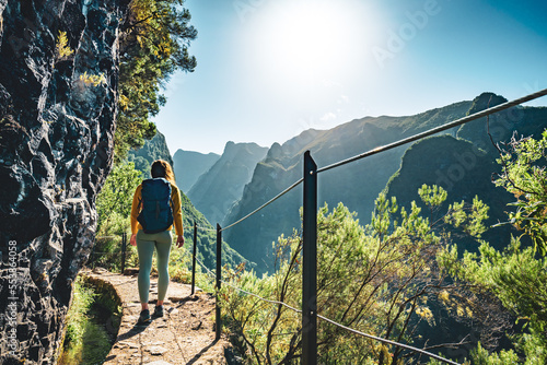 Backpacker woman walking along sunny hiking trail below large rock wall along water channel at steep cliff through Madeira's rainforest. Levada of Caldeirão Verde, Madeira Island, Portugal, Europe.