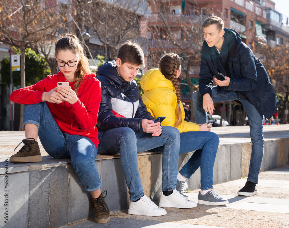 Fototapeta premium Portrait of modern teenagers hanging out on streets of city on warm spring day, using phones and chatting