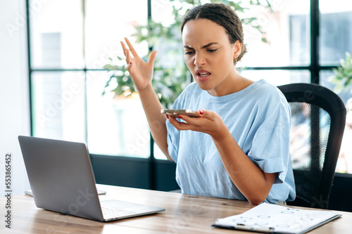 Annoyed angry young brazilian or latino business lady, company director, supervisor, yells irritably while speakerphone conversation, gesturing with hand, annoyed by poor performance of employees photo