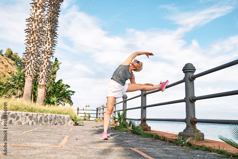 Young beautiful sportive woman training over seaside promenade, stretching legs before jogging. Fitness, sport and healthy lifestyle concept.