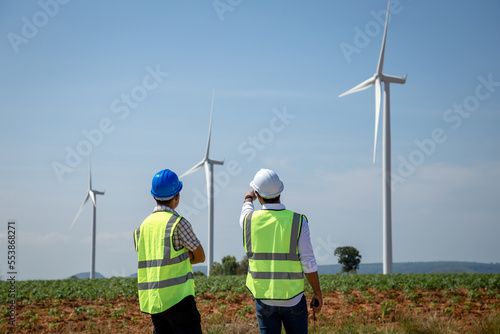 Two engineers wearing uniform and safety helmet work in wind turbine farm. Asian people engineers working at renewable energy farm