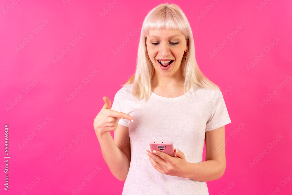 Blonde caucasian girl on pink studio background, copy space, pointing at mobile phone Blonde caucasian girl on pink studio background, copy space, pointing at mobile phone