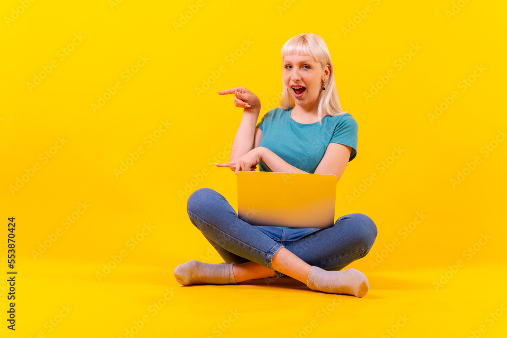 Sitting with a laptop pointing to the left, blonde caucasian girl in studio on yellow background Sitting with a laptop pointing to the left, blonde caucasian girl in studio on yellow background