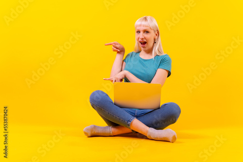 Sitting with a laptop pointing to the left, blonde caucasian girl in studio on yellow background © unai