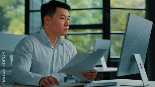 Tired unhappy Asian man worker manager sit at office doing routine paperwork read contract documents budget invoice pay bills online analyze business plan check documentation professional paper audit