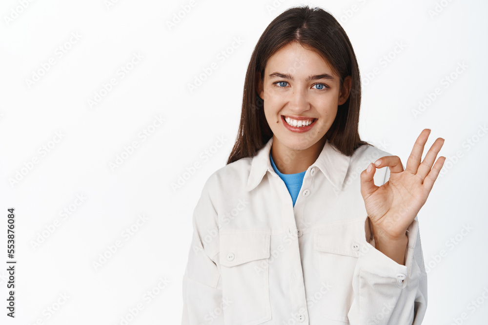 Close up of smiling beautiful young woman, female student showing praise, compliment, okay sign, standing over white background Close up of smiling beautiful young woman, female student showing praise, compliment, okay sign, standing over white background