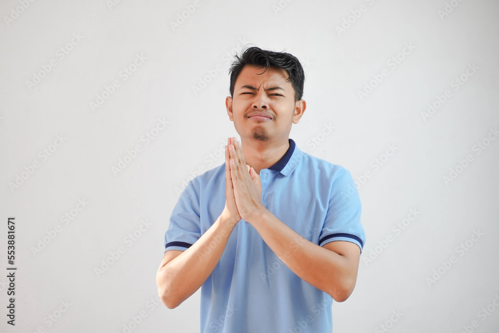 Young asian man wearing casual blue t shirt over white background begging with hands together with hope expression on face very emotional and worried. begging. sad face Young asian man wearing casual blue t shirt over white background begging with hands together with hope expression on face very emotional and worried. begging. sad face