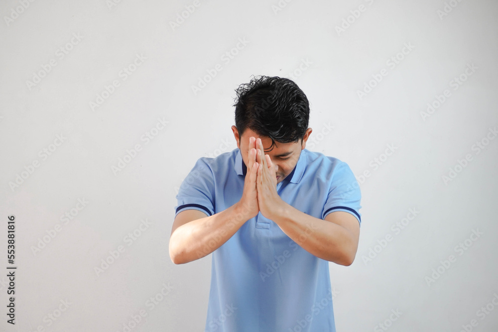 Young asian man wearing casual blue t shirt over white background begging with hands together with hope expression on face very emotional and worried. begging. sad face Young asian man wearing casual blue t shirt over white background begging with hands together with hope expression on face very emotional and worried. begging. sad face