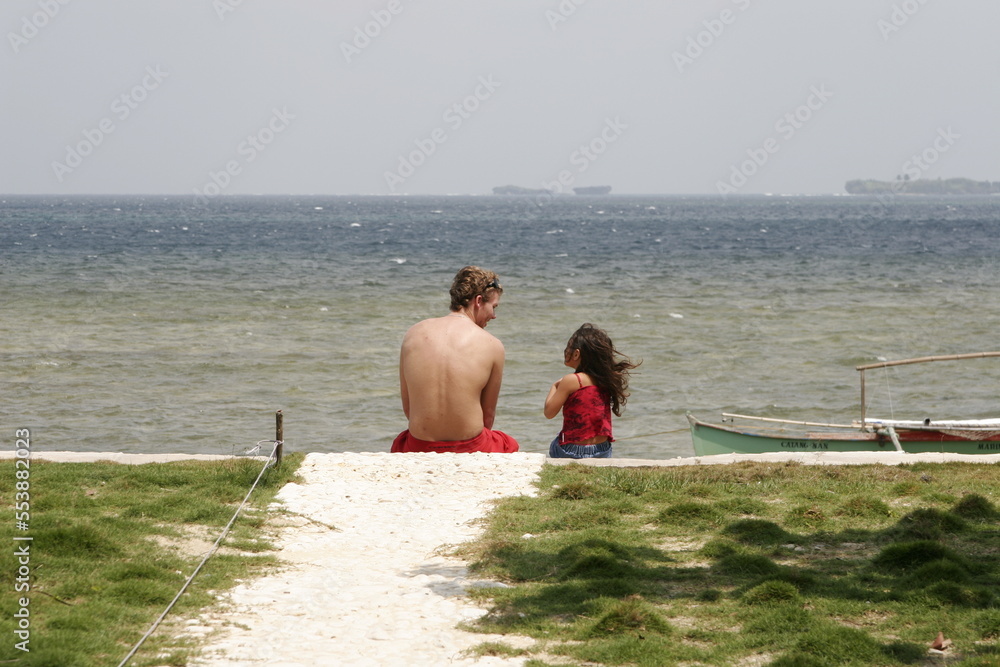 Father and daughter with backs turned, sitting at the beach on a sunny day