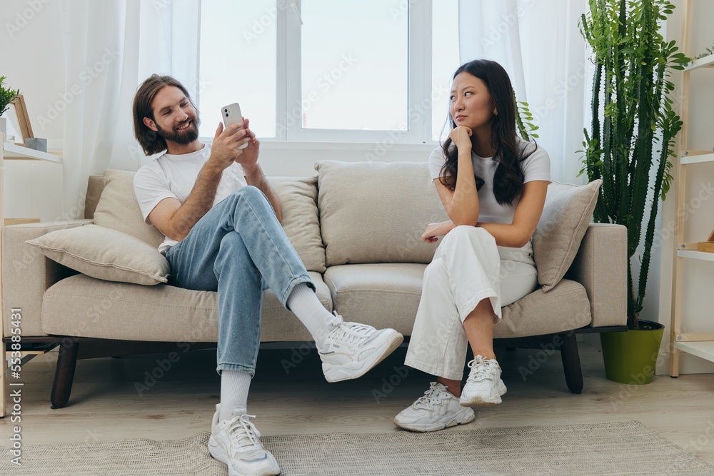 A man looks at his phone screen and smiles during an argument with his girlfriend. The angry and hurt woman looks in his direction and is sad. Family discord at home, phone addiction