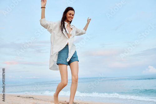 Happy tanned woman in white swimsuit shirt and denim shorts walks on the beach on the sand by the ocean with wet hair after swimming, sunset light and pink clouds in Bali