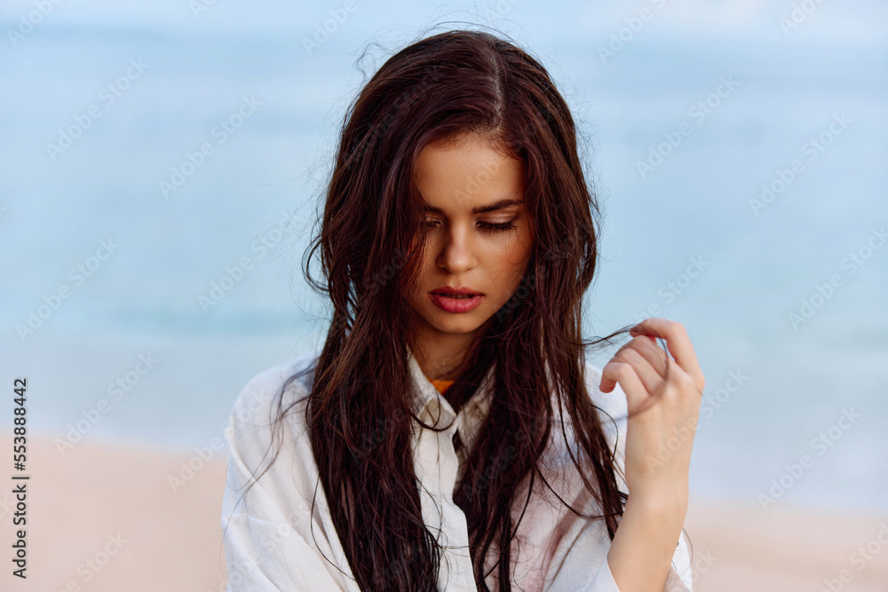Portrait of a beautiful pensive woman with tanned skin in a yellow tank top and white beach shirt with wet hair after swimming on the ocean beach sunset light